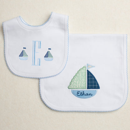 Set of two white cotton knit items with a blue picot edge.  One is a burp pad with a hand appliqued sailboat that is customized with an embroidered name.  The other piece is a bib with a monogram and two mini embroidered sailboats.