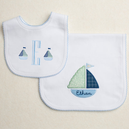 Set of two white cotton knit items with a blue picot edge.  One is a burp pad with a hand appliqued sailboat that is customized with an embroidered name.  The other piece is a bib with a monogram and two mini embroidered sailboats.