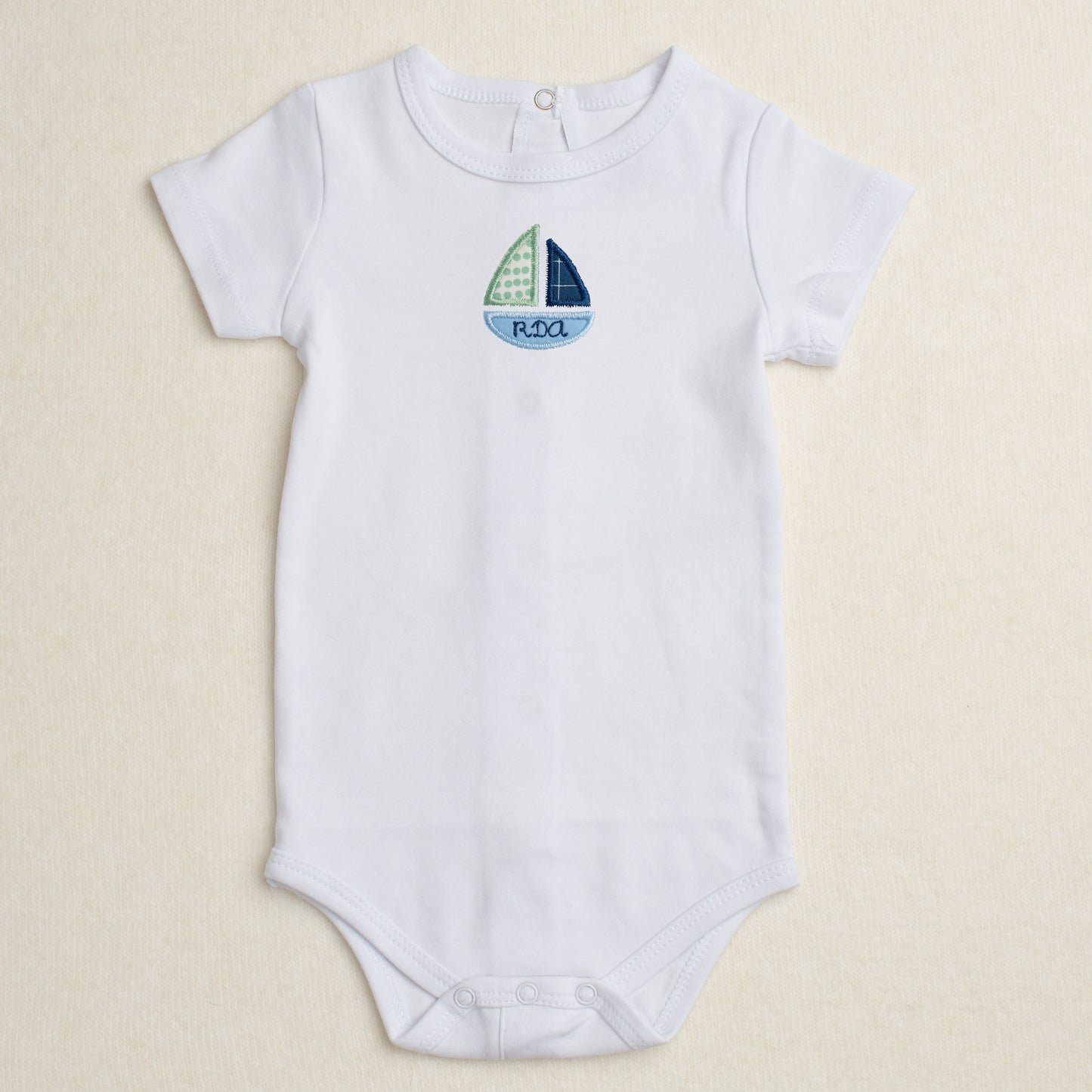 All cotton bodysuit with short sleeves and a blue sailboat applique that can be personalized with embroidery
