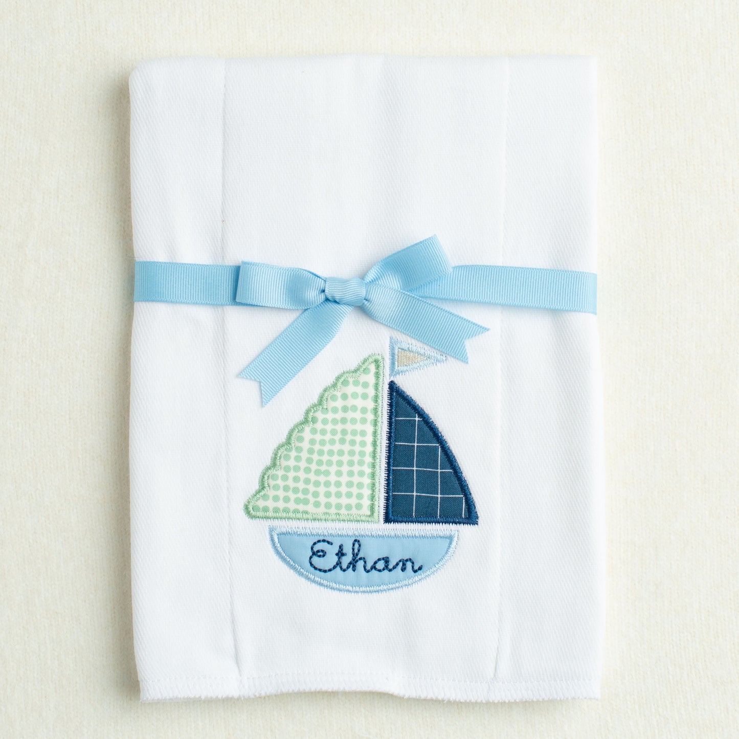 White burp pad with sailboat design and blue ribbon on a beige background
