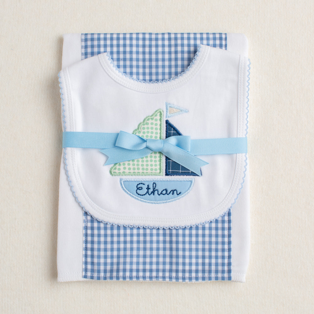 Handmade 2 piece gift set presented ready for gift giving folded and tied with a grosgrain ribbon.  Set includes blue check fabric burp pad along with a white bib with Deacon Lane's appliqued sailboat motif with a name custom embroidered in the bottom of the boat.