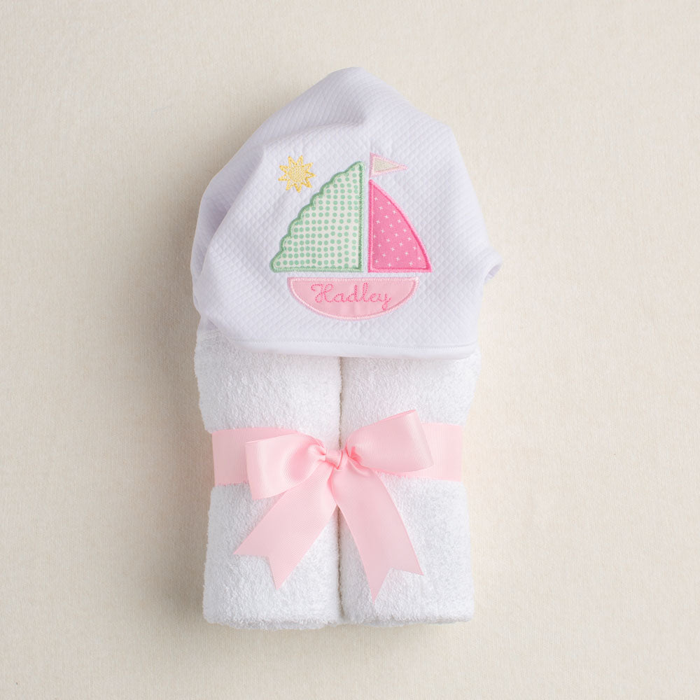 White hooded towel with pink sailboat design and pink ribbon on a beige background