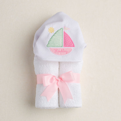 White hooded towel with pink sailboat design and pink ribbon on a beige background