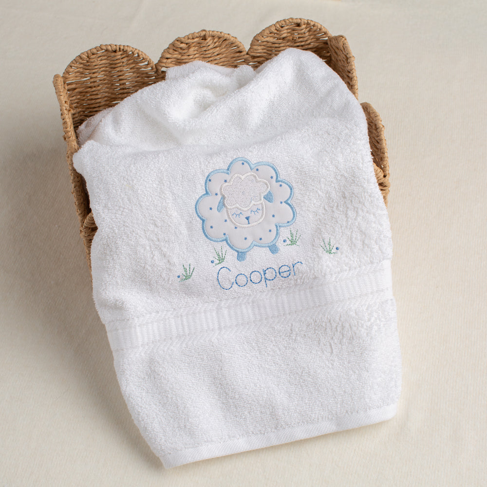 White towel with embroidered sheep design and 'Cooper' text, placed in a woven basket on a beige background.
