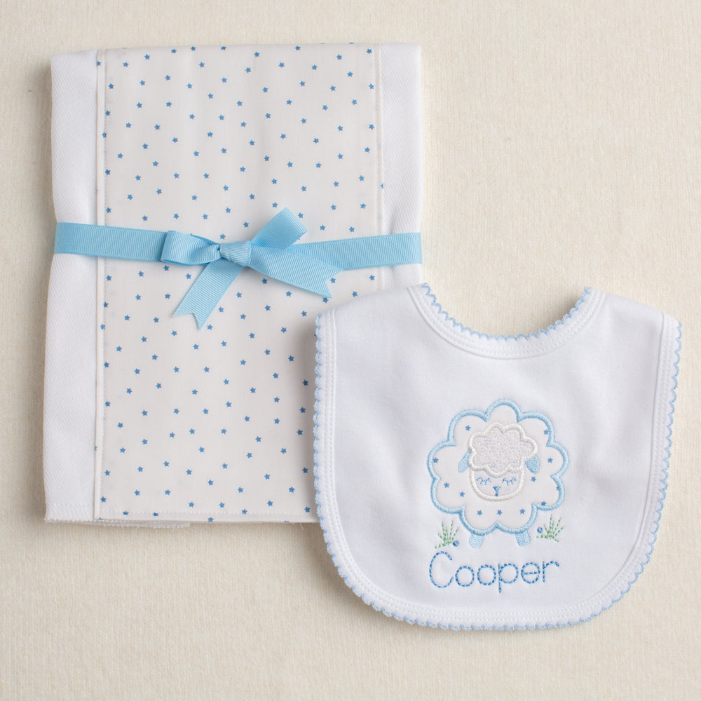 Baby bib with embroidered design and name 'Cooper' next to a folded burp pad with a blue ribbon on a beige background