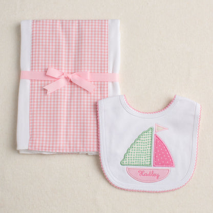 Two piece set showing both pieces of the gift - one is an appliqued sailboat bib in pink and the other is a fabric burp cloth with a pink gingham panel.