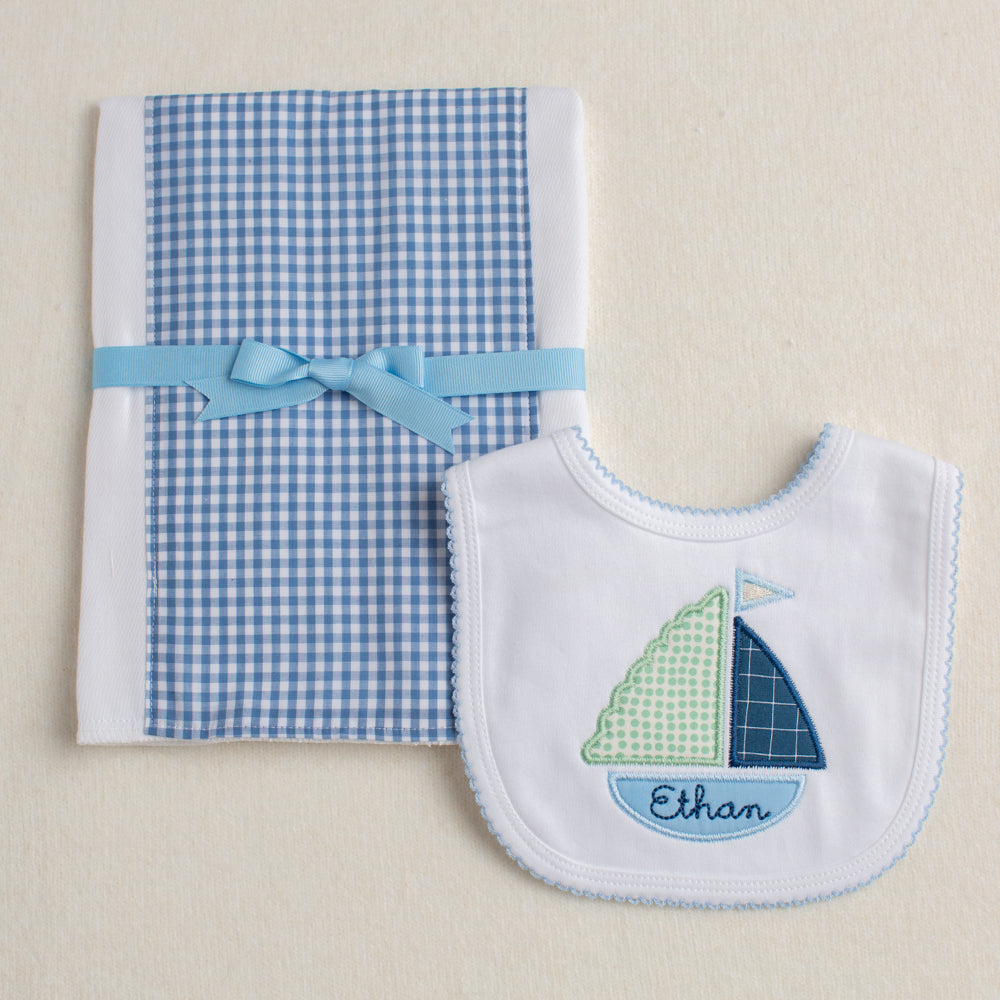 2 piece boutique baby gift set with a blue check fabric burp pad and a white bib with blue picot edging appliqued with a beautiful green and blue boat with a custom name embroidered in the bottom of the boat.
