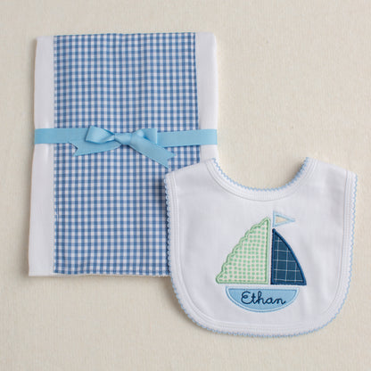 2 piece boutique baby gift set with a blue check fabric burp pad and a white bib with blue picot edging appliqued with a beautiful green and blue boat with a custom name embroidered in the bottom of the boat.
