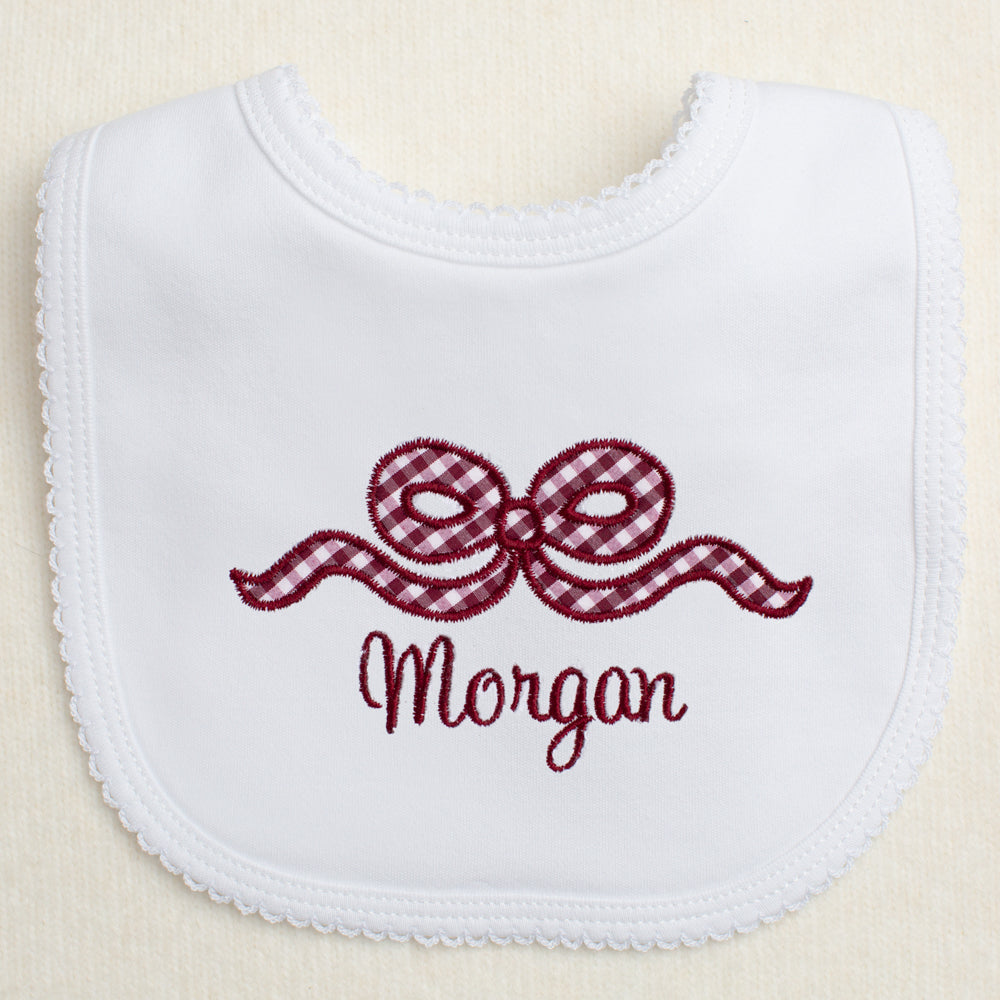 white bib with a picot edge with an appliqued maroon bow and a personalized, embroidered name for Texas A + M fans