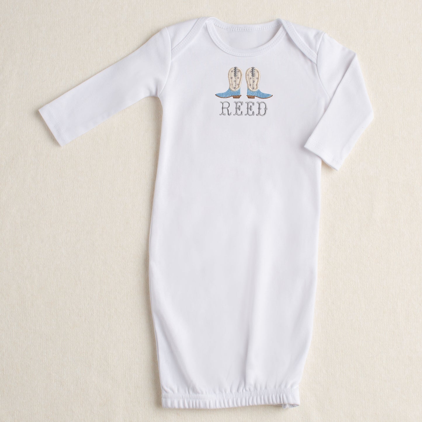 White baby gown with a pair of handmade applique cowboy boots in shades of light blue, cream, and grey. A Deacon Lane original design customized with an embroidered name in a Western Fishtail font.
