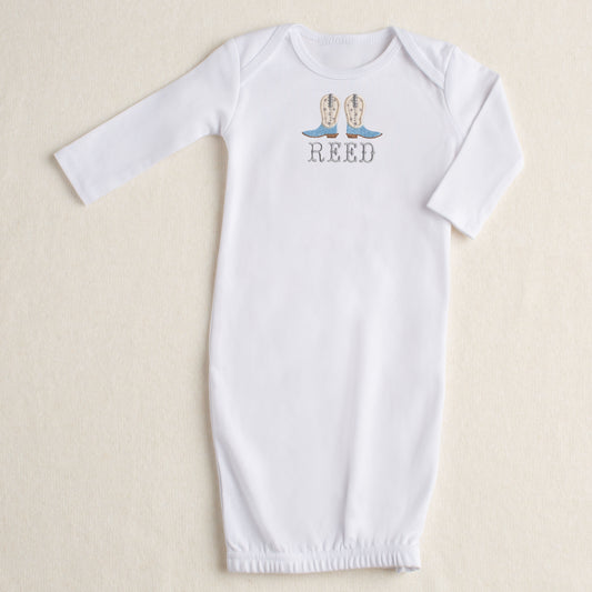 White baby gown with a pair of handmade applique cowboy boots in shades of light blue, cream, and grey. A Deacon Lane original design customized with an embroidered name in a Western Fishtail font.
