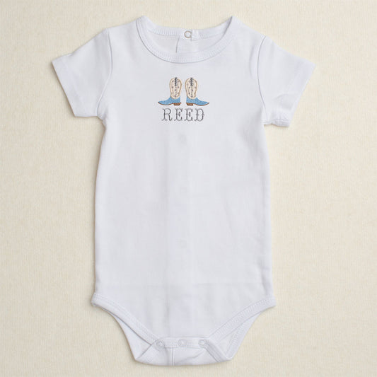 White baby onesie with appliqued cowboy boots and the name 'REED' embroidered underneath in a western drop-shadow font .