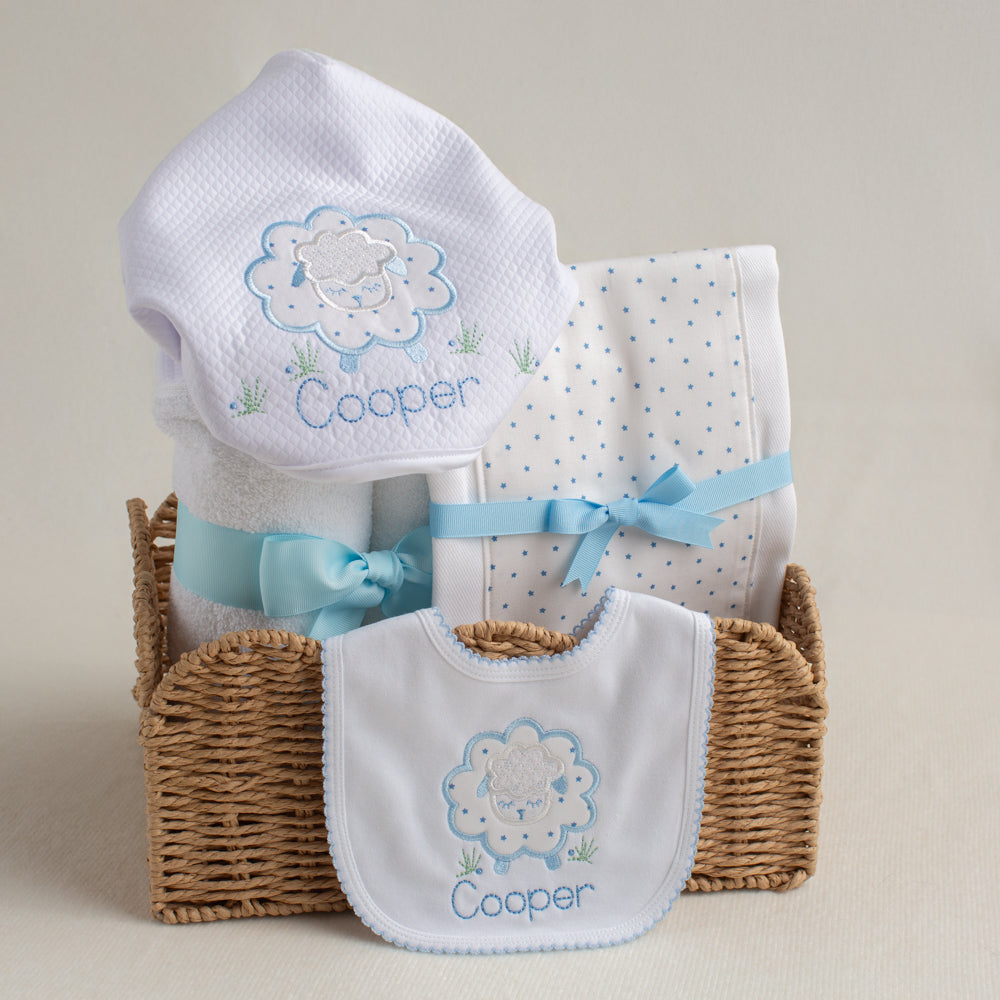 Baby gift set with white towel, burp pad and bib in a blue applique sheep design  in a basket on a beige background