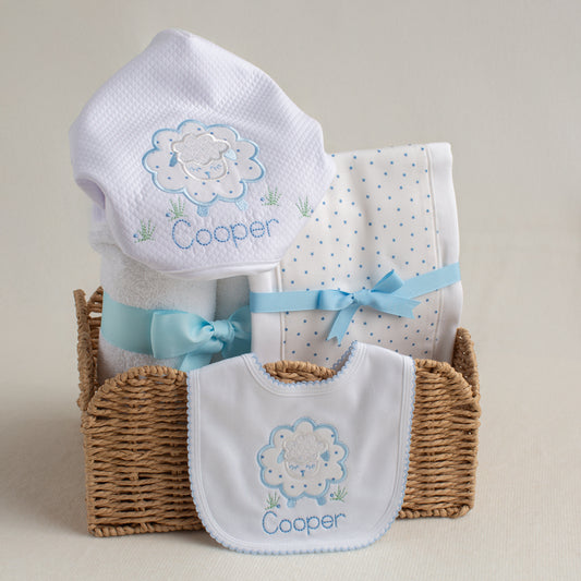 Baby gift set with white towel, burp pad and bib in a blue applique sheep design  in a basket on a beige background