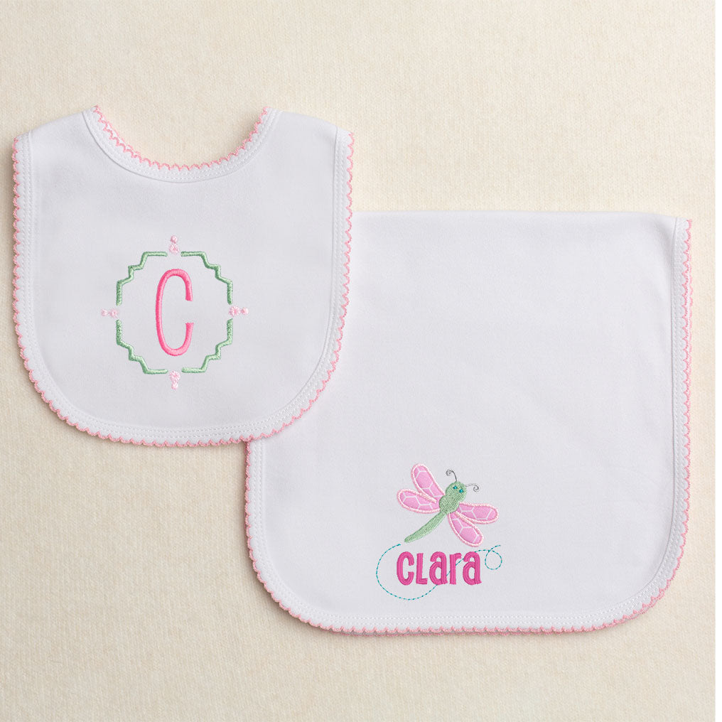 Custom baby gift set featuring two white knit items with a pink picot edge.  The bib had a single monogram initial surrounded by an intricate frame and the burp pad has an appliqued pink dragonfly with a personalized name