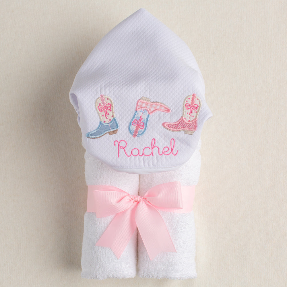 White hooded towel with embroidered cowboy boots and the name 'Rachel', tied with a pink ribbon on a beige background.
