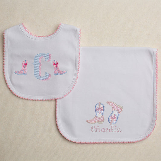 Monogrammed cotton bib with pink picot trim featuring a western theme font with cowboy boots with bows.  Set includes a personalized burp pad with pink picot trim with appliqued cowboy boots