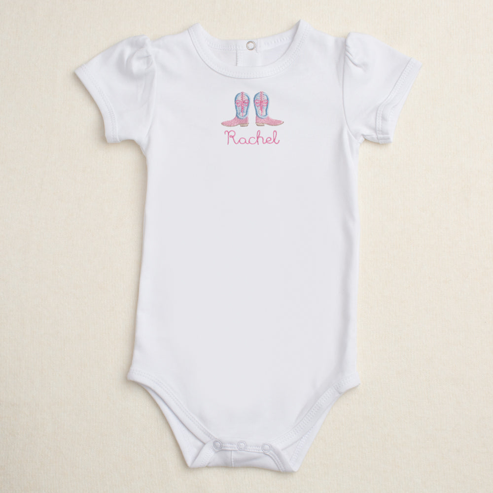 White baby onesie with Shabby Chic cowgirl boots appliqued and personalized with the name 'Rachel' on a beige background