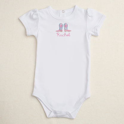 White baby onesie with Shabby Chic cowgirl boots appliqued and personalized with the name 'Rachel' on a beige background