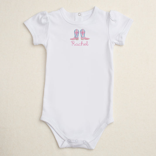 White baby onesie with Shabby Chic cowgirl boots appliqued and personalized with the name 'Rachel' on a beige background