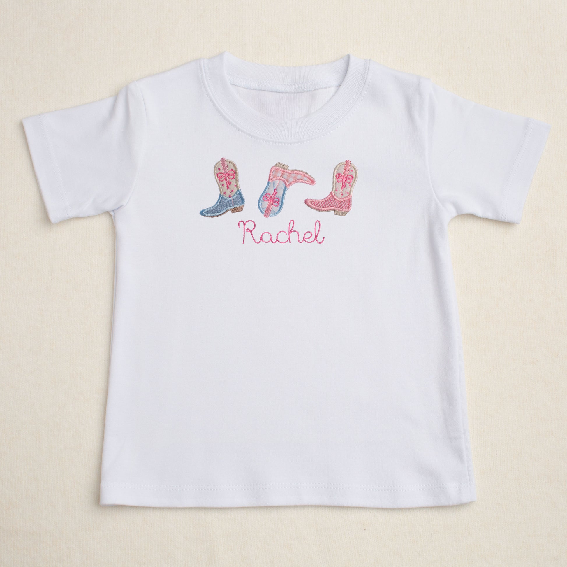 White personalized t-shirt with embroidered pink cowboy boots and the name 'Rachel' on a beige background