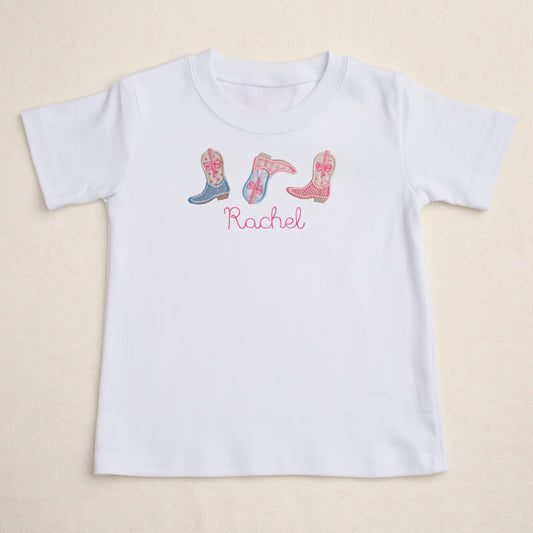 White personalized t-shirt with embroidered pink cowboy boots and the name 'Rachel' on a beige background